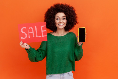 满意的微笑女人，非洲发型，穿着绿色休闲风格毛衣，拿着销售卡和带空白黑色显示屏的智能手机。