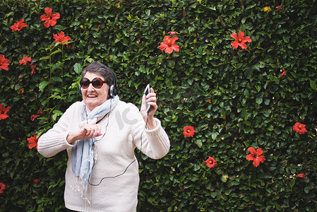 有趣的老妇人戴着耳机在智能手机上跳舞听音乐，微笑着在带花墙的花园里享受庆祝退休的乐趣