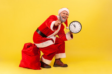 快乐积极的老人，留着灰胡子，穿着圣诞老人服装，坐在红色袋子上，手里拿着礼物，手里拿着挂钟，指着相机。