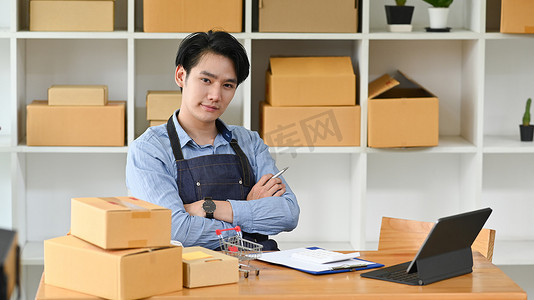 自信的男子小企业主穿着围裙坐在纸板箱中间，看着相机。
