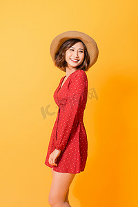 连衣裙亚洲女性摄影照片_橙色背景中身穿红色连衣裙、头戴草帽、快乐、精力充沛的亚洲女性的摄影棚照片