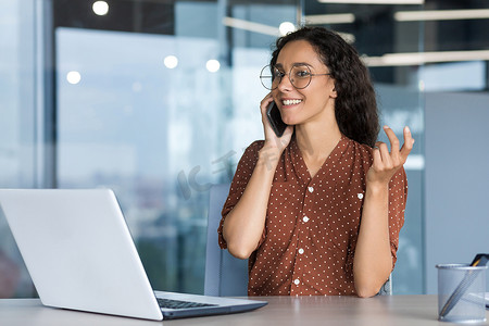 西班牙女摄影照片_年轻成功的西班牙裔女商人在现代办公室工作，女员工高兴地与同事通过电话交谈，微笑着，坐在办公桌前使用笔记本电脑