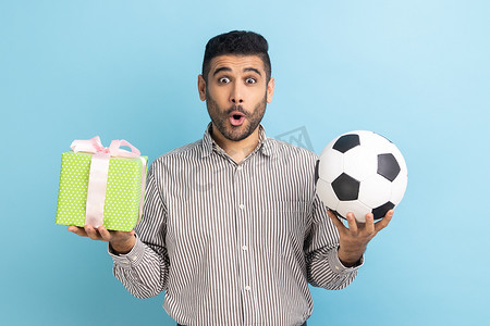 端午节礼摄影照片_惊讶的商人站着，惊讶地看着镜头，手里拿着足球和礼物盒