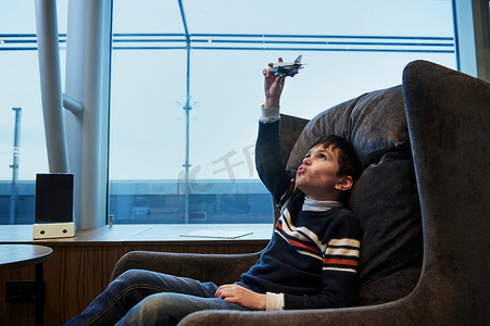 在国际机场出发航站楼等待登机时，十几岁的男孩坐在窗边玩玩具飞机，俯瞰跑道