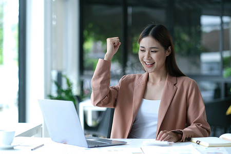 在现代化的办公室里，年轻的亚洲女商人在工作中用笔记本电脑开心地微笑着，表现出成功的喜悦。