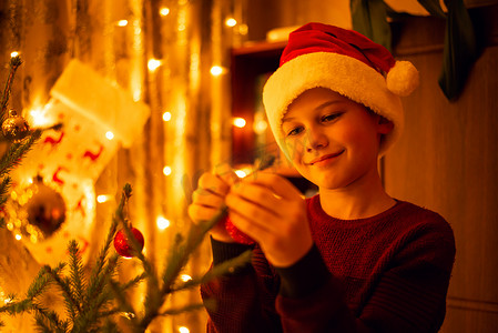 戴着圣诞帽的微笑男孩装饰着房间里的冷杉美景，里面充满了花环的黄色灯光
