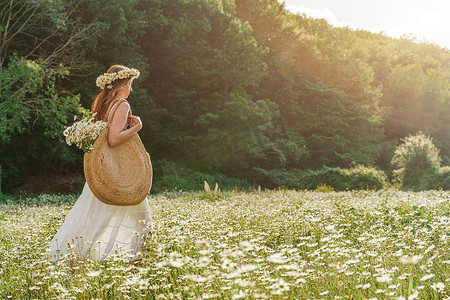 一位身穿白色背心裙、肩上挎着一个大柳条袋的雏菊花环的年轻女子正在一片雏菊田中漫步，背景是森林和夕阳的光芒。