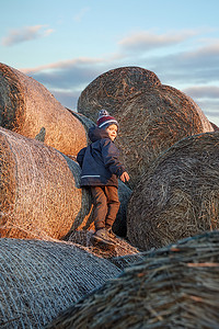 秋天的傍晚，小男孩穿着秋装，戴着祖母的针织帽，在外面一堆柔软的稻草堆上玩耍