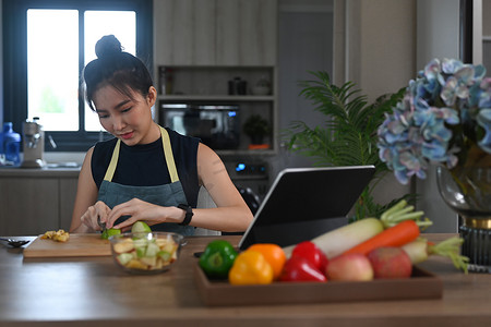 丰富的数字摄影照片_穿着围裙的美丽亚洲女人准备健康沙拉，并在厨房的数字平板电脑上阅读在线食谱。