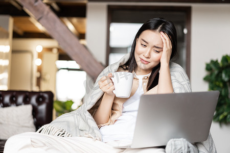 年轻患病的亚洲女孩待在家里隔离、感染流感或科维德、在笔记本电脑上观看视频和喝茶