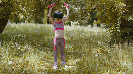 戴着 VR 耳机头盔的运动儿童在公园户外用哑铃进行健身锻炼