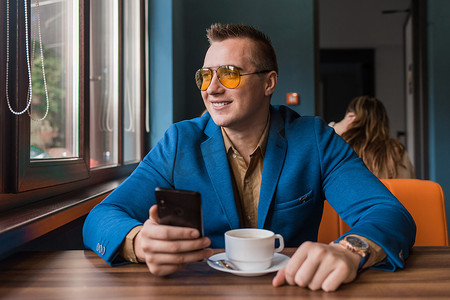 望向窗外摄影照片_一位戴着墨镜、夹克和衬衫、面带微笑的欧洲商人坐在咖啡馆的桌子旁喝咖啡休息，手里拿着智能手机或手机，望向窗外