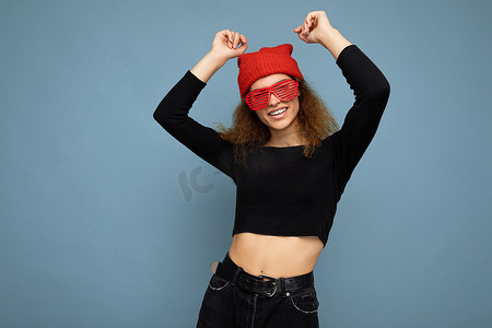 照片中，漂亮迷人的年轻卷发深色金发女郎孤立地站在蓝色背景墙上，上面有文字复制空间，穿着日常时尚的黑色上衣，戴着红色的帽子和眼镜。