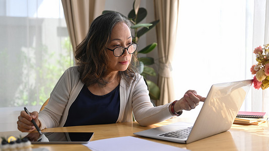 集中精力的成熟女商人在家工作,坐在现代家里,在笔记本电脑上进行在线