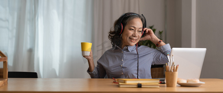 戴着耳机的快乐亚洲老年女性在舒适的家客厅里用笔记本电脑听歌