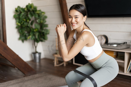 可爱的亚洲健身女孩在家锻炼，腿上拉着弹力绳蹲下，锻炼身体健康