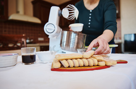 马斯卡彭摄影照片_在舒适的家庭木制厨房里准备自制意大利甜点提拉米苏时，用海绵饼干手指软专注于女手