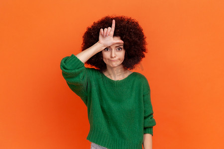 悲伤的女人，留着非洲发型，穿着绿色休闲风格的毛衣，在额头上做出失败者的手势，看上去脾气暴躁，对失败感到沮丧。