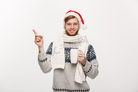 圣诞节概念 — 穿着毛衣和圣诞老人​​帽子的年轻胡子男子拿着一个热咖啡杯，手指向一侧，与白色隔离，有复制空间。