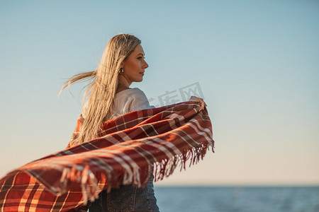 迷人的金发白种女人在日落时在海滩上享受时光，裹着毯子走着，看着旁边，背景是夕阳的天空和大海。