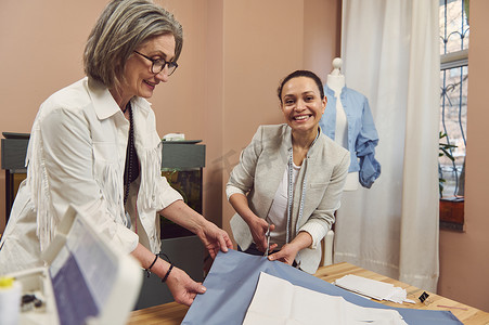 两位成熟女裁缝在时装设计工作室缝制经典女式衬衫时在布料上制作衣服图案的齐腰肖像