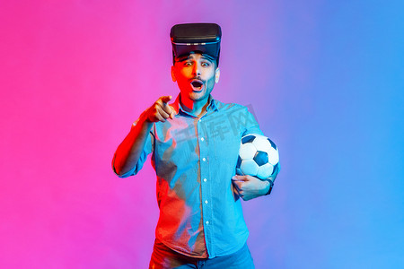 戴着虚拟现实眼镜的男子，与足球合影，手指指向相机。