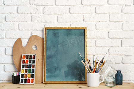 相框作为画架，木桌上有艺术家的工具，靠在白砖墙上