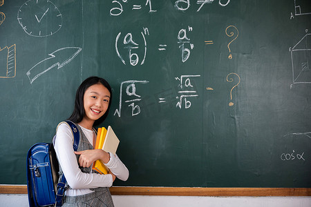 快乐美丽的亚洲女学生拿着书站在教室黑板前
