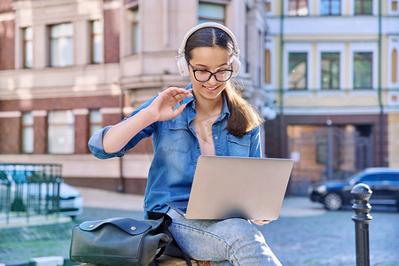 戴耳机的少女学生在户外、城市背景下使用笔记本电脑
