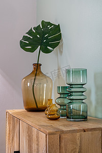 透明的黄色和绿色花瓶，用于鲜花和大瓶，木制橱柜上有龟背竹叶。