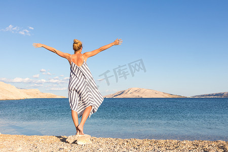 快乐无忧无虑的女人举起双臂，穿着漂亮的条纹夏装，在克罗地亚帕格岛的白色卵石海滩上享受午后时光