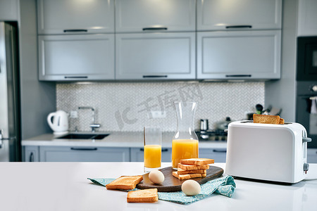 面包鸡蛋橙汁摄影照片_浅色厨房桌上的烤面包机，配有新鲜面包、鸡蛋和一杯橙汁