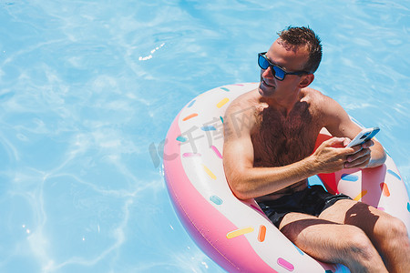 手机夏季夏季摄影照片_一个有魅力的男人正在游泳池的充气环上放松。