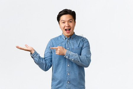 微笑开朗可爱的亚洲男子发布公告，展示手头的新产品，用手指指着广告的空白处，站在白色背景中逗乐