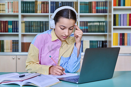 戴着耳机的高中生女孩在学校图书馆使用书籍和笔记本电脑学习