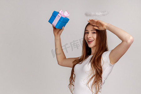 端午节礼摄影照片_年轻友好的女人拿着蓝色包装的礼品盒，看着远处，对着镜头微笑的肖像
