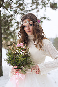 冰雪婚礼摄影照片_美丽的新娘穿着白色连衣裙，在冰雪覆盖的冬季森林里捧着花束。