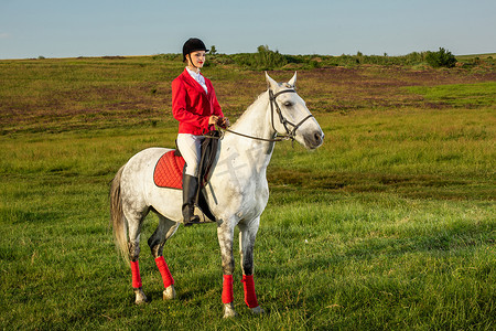 女骑手摄影照片_年轻的女骑手，穿着红色的 redingote 和白色的马裤，她的马在夕阳的余晖下。