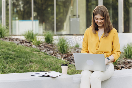 专注于欧洲女商人的企业家在电脑上打字，用笔记本电脑学习在线课程，微笑的年轻女孩在办公室外远程工作，使用电脑设备坐在长凳上