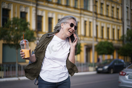 快乐的成熟欧洲女性笑着打电话享受下班后的空闲时间，或者在城市背景下用塑料杯在旅途中喝果汁。