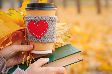 秋意浓情摄影照片_一本书、一束秋叶和一个手里拿着咖啡的纸杯，户外的秋意