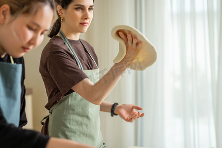 厨师女厨师摄影照片_两位年轻的女厨师在厨房里煮披萨，其中一位在手里捻着卷好的面团