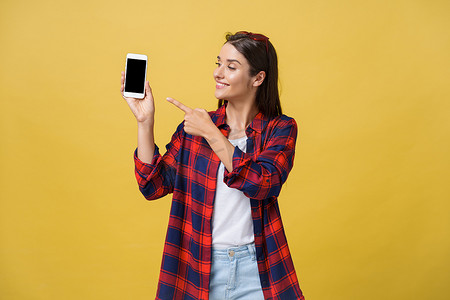 手机夏季夏季摄影照片_一个穿着夏季服装的快乐年轻女孩的肖像，手指着黄色背景中突显的空白屏幕手机