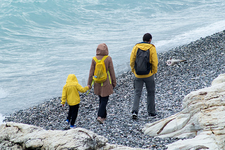 2022岩石摄影照片_俄罗斯索契 — 2022年5月9日：一个男人、一个女人和一个孩子穿着黄色雨衣沿着卵石滩散步