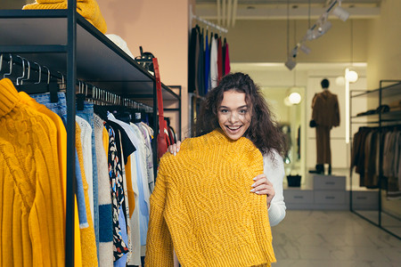 一位年轻漂亮的卷发女子在服装店商场寻找一件温暖的黄色夹克，在柜台上的衣架上检查它。