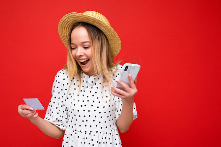 一张漂亮的正面照片让身穿夏装的年轻金发女子使用手机并持有信用卡在红色背景下在线支付感到惊讶。
