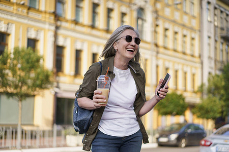 欧洲杯券摄影照片_头发花白的欧洲高级女性很高兴享受下班后或旅行后的空闲时间，一边拿着手机，一边在城市背景下使用塑料杯喝果汁。