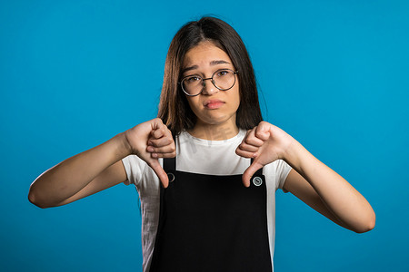 年轻的亚洲女孩站在蓝色工作室背景上表达不满，并对着镜头表现出拇指向下的手势。