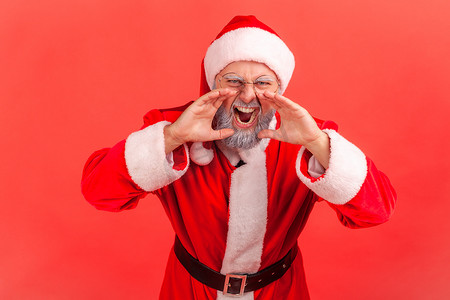 喊叫表情摄影照片_身穿圣诞老人服装、留着灰胡子的老人的肖像，脸上表情愤怒，双手靠近嘴部尖叫。