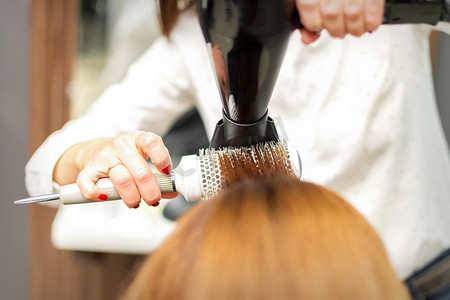 长头发短头发摄影照片_一位专业美发师正在用吹风机和圆刷烘干长红头发，特写。